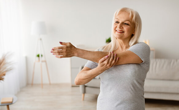 Domestic Sports Concept. Athletic mature woman in sportswear doing fitness exercises during covid quarantine, free space. Flexible senior lady stretching her arm, warming up before home workout
