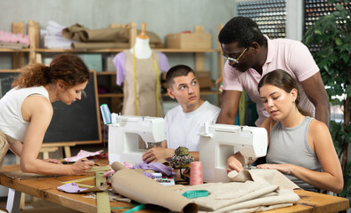 During lesson, African male teacher helps student sew, while others sew and work with paper, draw dress pattern. Interest group, activities, hobbies, vocational training..