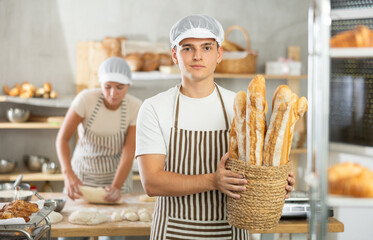 Young man are happy with large amount of made bread, family bakery, storage room. Guy participant of bread production holds basket of baguettes, successful entrepreneurs in back room of bakery