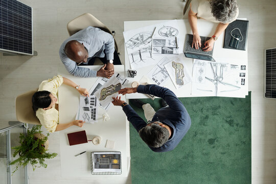 Diverse group of middle aged professionals collaborating on architectural project, reviewing blueprints and discussing plans around table, one using laptop, top view