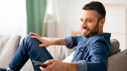 Relaxed bearded man switching channels on TV at home, holding TV remote, smiling middle-aged man sitting on sofa in living room, watching television, enjoying lazy weekend, side view, copy space