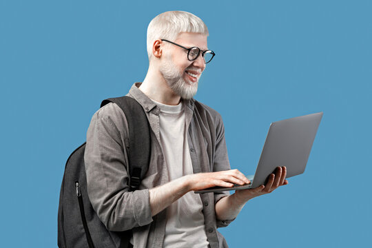 Cool guy with unusual appearance using laptop for online work or studies, wearing backpack over turquoise studio background. Happy young man communicating on web, browsing internet