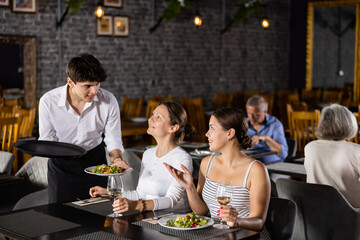 Young male waiter in uniform serves ordered dish to two women in restaurant..