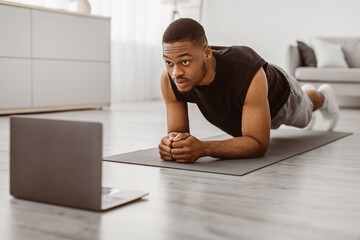 Fototapeta premium Workout. African American Guy Doing Elbow Plank Exercise Training At Laptop Computer In Living Room At Home. Online Fitness And Sporty Lifestyle, Bodybuilding Concept. Selective Focus