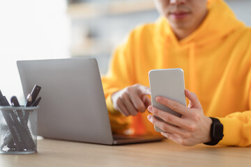 People And Technology. Closeup cropped view of unrecognizable guy in bright yellow hoodie holding smartphone using laptop computer, reading business text message, sitting at desk, selective focus
