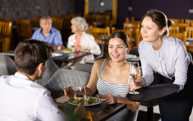Young male and female colleagues have fun chatting and spending time in restaurant after working day. Female waitress serves order of wine and wishes cafe visitors pleasant appetite and good time