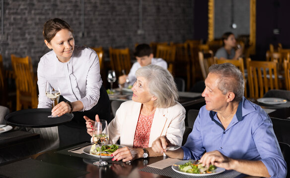Young female waiter serves mature couple of positive spouses in restaurant. Hostess serves puts on table order for dinner dishes of national Mediterranean cuisine