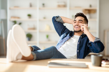 Obraz premium Relaxed arab man freelancer having short break while working on laptop from home, sitting at workdesk with legs up and leaning back, smiling and having phone conversation, looking at copy space