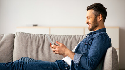 Happy middle-aged man in casual resting on couch at home, using modern smartphone, copy space....
