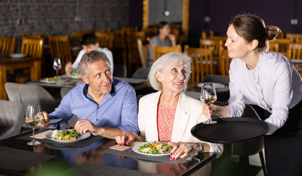 Elderly spouses sitting next to each other in cozy cafe, drinking wine and eating light salad snack. Waitress girl restaurateur serves customers plate with dinner order
