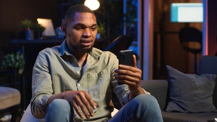 African american guy attends web conference call on laptop sitting at home, joining an remote webinar to discuss about solving daily tasks. Teleconference in the living room. Camera B.