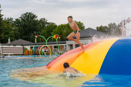 Cheerful boy in swim shorts jumping off inflatable water trampoline into outdoor pool at aqua park on sunny summer day. Concept of childhood joy, active holiday, water fun, adventure and recreation.