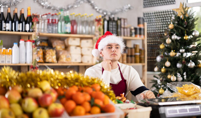 Male salesman in a Santa hat sits thoughtfully behind a counter with fruits and scales. Salesman in a supermarket alone celebrates the New Year