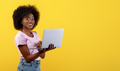 Online work. Excited black woman holding and using newest laptop computer, standing over yellow studio background with copy space. Cheerful female typing on keyboard and smiling to camera