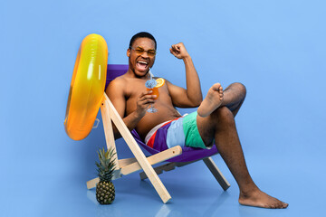 Full length of excited black man drinking tropical cocktail in lounge chair, gesturing YES over blue background. Young African American guy chilling on beach during summer vacation
