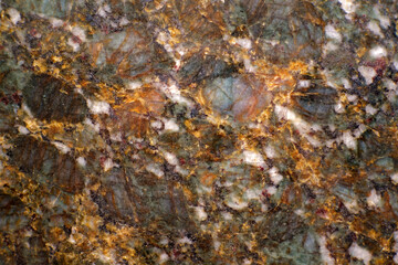 Close-up of natural granite stone surface showing multicolored mineral patterns. The detailed texture with green, gold and white tones highlights the beauty of geology and decorative stone design.