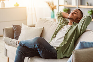 Obraz premium Relaxation And Comfort. Contented Black Guy Relaxing With Eyes Closed Holding Hands Behind Head Sitting On Sofa At Home. Man Enjoying Lazy Weekend In Living Room. Side View
