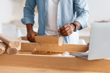 Black man screwing bolt to wooden table leg and using laptop at home interior. Handyman reading blog about repair furniture and carpentry, watching online instructions