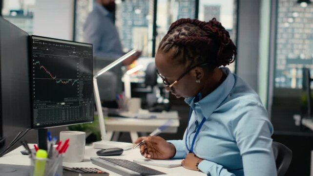 Equity analyst in prop firm office reviews quarterly earnings to assess future stock market value, taking notes. Trader conducts financial analysis to improve investments, writing down info, camera B