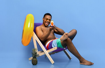 Playful black man relaxing in lounge chair with water gun, pointing toy pistol at camera over blue studio background, full length. Funny young guy enjoying his beach holiday