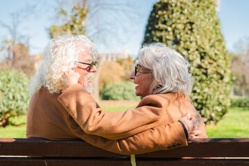 Happy senior couple sharing a loving embrace and laughter while seated together on a bench, enjoying a special romantic moment that highlights their bond and affection for each other in a serene park
