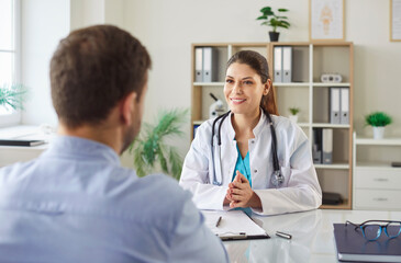 Friendly female doctor sitting at her desk with male patient. Smiling young woman physician or...