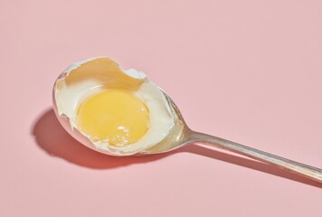 Close-up of a perfectly cooked sunny-side-up egg with a runny yolk on a stainless steel spoon against a soft pink background