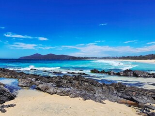 Scenic Coastal Beach with Rocky Shoreline and Turquoise Waves crashing under Clear Blue Sky, Mountains in Background
