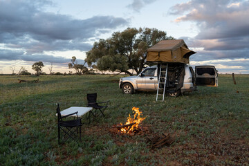 A four-wheel drive with a rooftop tent and an open back door is parked on a grassy field. A small fire burns in front of the vehicle, next to a table and two camping chairs. © pauline.mongarny
