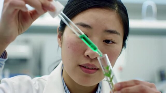 A focused female scientist in a white lab coat meticulously adds a green chemical from a pipette to a test tube containing a pink substance during an experiment.