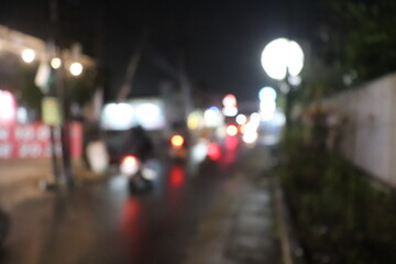 Out-of-focus city street at night, filled with blurred car headlights, glowing taillights, and scattered streetlights that create a dreamy bokeh effect. The image reflects the hustle of nightlife