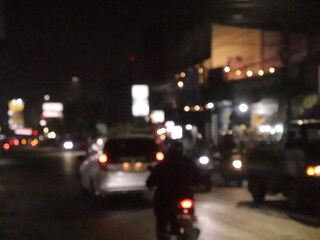Out-of-focus city street at night, filled with blurred car headlights, glowing taillights, and scattered streetlights that create a dreamy bokeh effect. The image reflects the hustle of nightlife