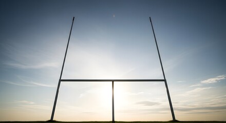 Silhouette of rugby goalposts against a bright, hazy blue sky at sunset