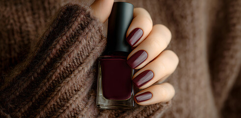 Close-up female hand with a purple manicure holding nail polish bottle, fashionable short square nails in scarlet tone, styled with a brown sweater in natural light for beauty and elegance concept