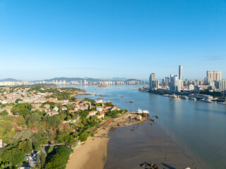 Fototapeta premium Xiamen China presents a stunning coastal panorama, where towering city structures meet the calm water and green shores under a bright, open sky.