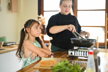 Upset child watching her mom cooking food in a frying pan, expressing impatience or disinterest