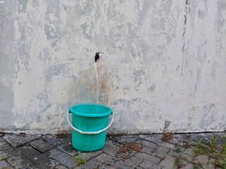 A green plastic bucket is placed directly under a water tap attached to the wall. The bucket looks empty, but water is flowing from the tap into the bucket.