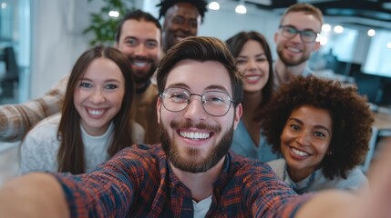 A group of workers gathers around a colleague whose work anniversary it is, taking a selfie to capture the joyful moment. The anniversary person is beaming, surrounded by smiling faces in a room fille