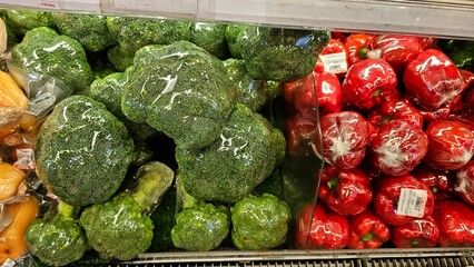 Close-up shot of several heads of broccoli and bell pepper, individually wrapped in plastic. They are arranged closely together, for sale in a grocery store