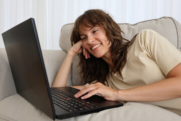 Fototapeta premium Beautiful smiling curly brunette lying on the sofa and working at the laptop at home