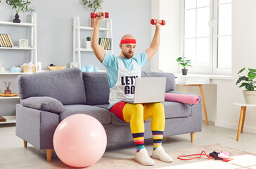 Funny man in extravagant colored sportswear sitting on sofa with laptop, doing exercises, lifting dumbbells. Weird concentrated sportsman following online video tutorial, having sport workout at home.
