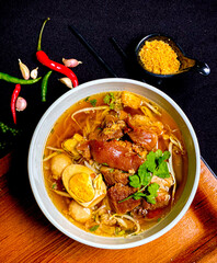 Thai braised pork leg noodle soup served in a bowl with fresh herbs, boiled egg, and green onions. Tender pork leg and rich savory broth with soft noodles, a traditional and appetizing Thai comfort fo