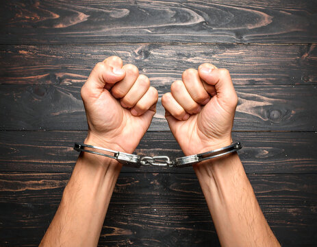 Close-up shot of human hands restrained by shiny metal handcuffs, on a wooden surface.