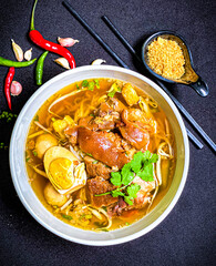 Thai braised pork leg noodle soup served in a bowl with fresh herbs, boiled egg, and green onions. Tender pork leg and rich savory broth with soft noodles, a traditional and appetizing Thai comfort fo