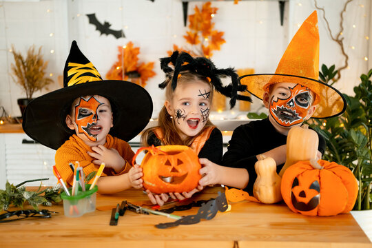 Halloween, kids boys and a girl in festive costumes and makeup are preparing for Halloween at home in the kitchen by coloring and carving pumpkins and scaring everyone - Powered by Adobe