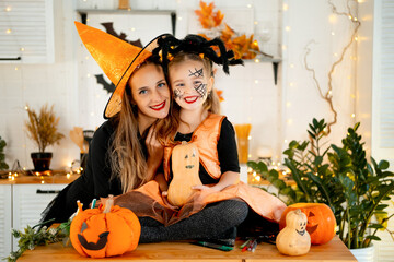 Halloween, a happy family a mother with a baby girl in festive costumes and makeup is preparing for Halloween at home in the kitchen by coloring and carving pumpkins and scaring everyone