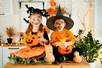 Halloween, two baby boy and girl in festive costumes and makeup are preparing for Halloween at home in the kitchen by coloring and carving pumpkins and scaring everyone