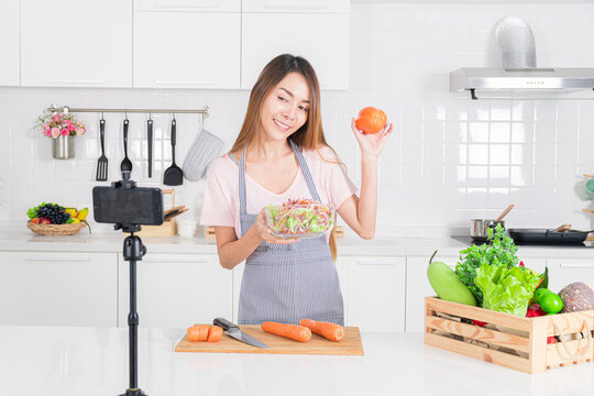 Engaging woman enthusiastically show freshly made salad and capsicum to her smartphone camera, creating content about healthy eating in her bright kitchen.