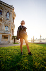 Fototapeta premium A stylish woman poses confidently on a grassy lawn with a historic building in the background during a warm sunset, creating a vibrant yet serene ambiance.