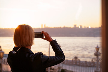 A woman enjoys a serene scenic view by capturing a beautiful sunset over the ocean using her smartphone, emphasizing tranquility and connection with nature.
